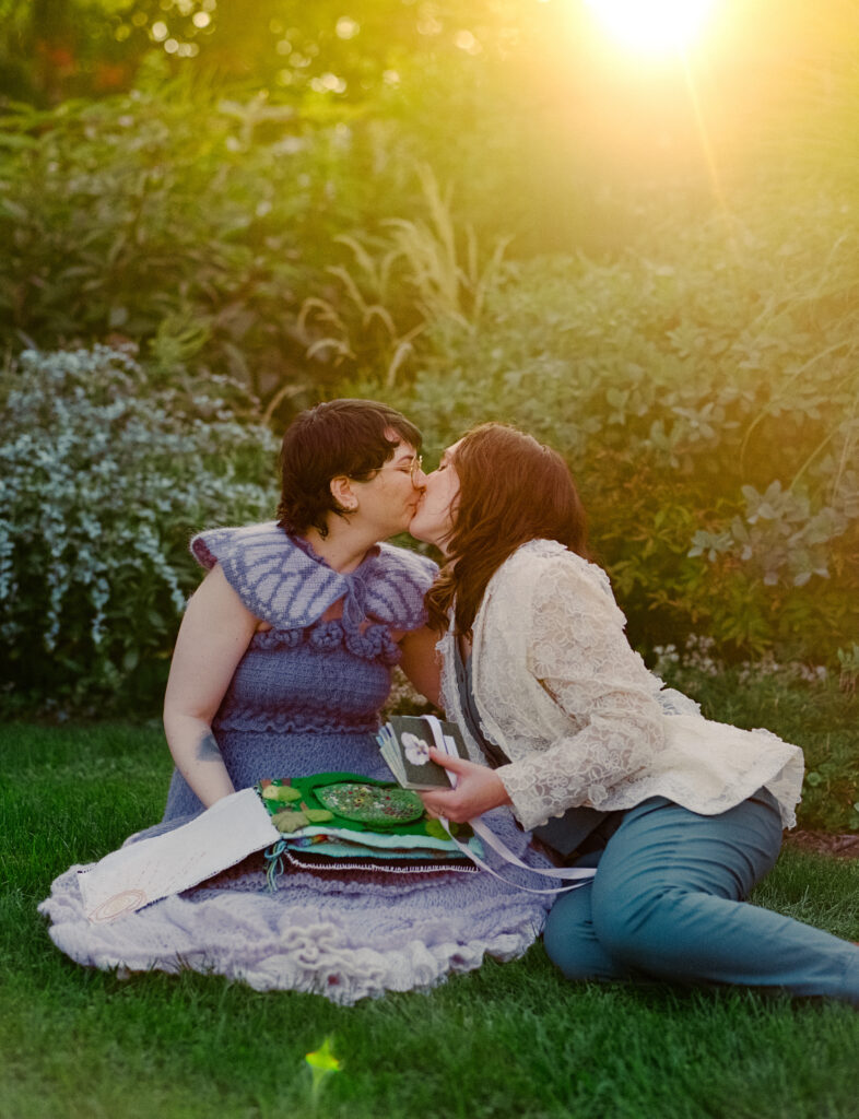 Newlyweds sit on the ground and lean in for a kiss, holding their gifts to each other in hand.