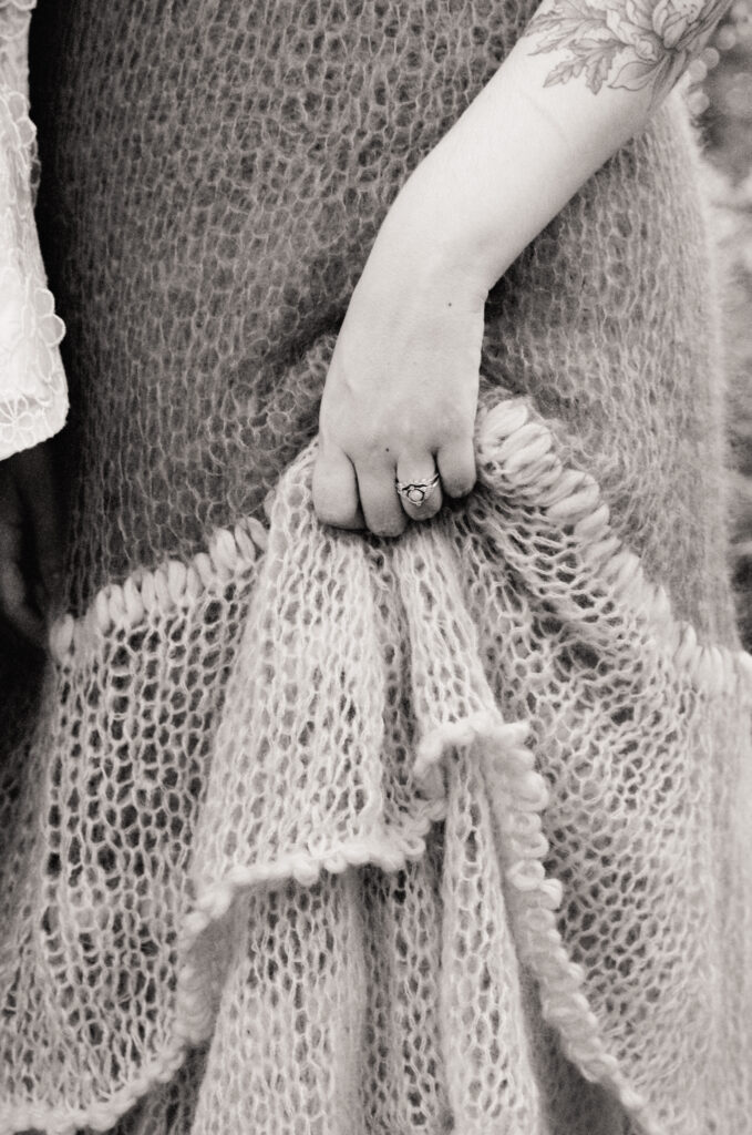 Black and white detail shot of crocheted dress being held up. The hand holding it showcases an engagement ring. 