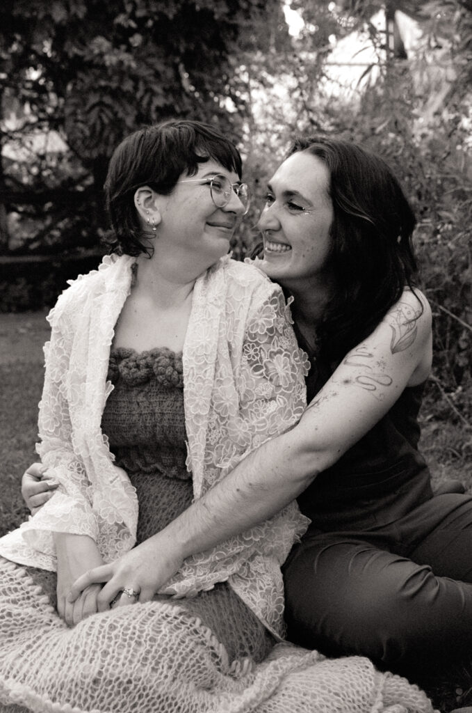 Black and white photograph of newlywed couple looking at each other and holding hands, sitting amongst tended gardens.