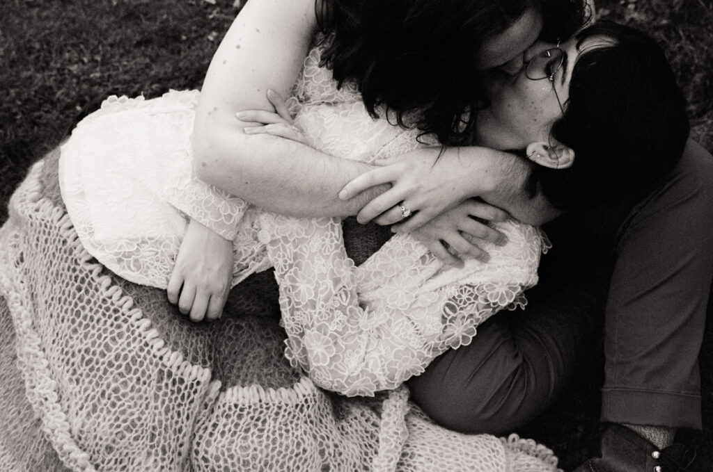 Black and white close up photograph  of newlywed couple laying on the ground and kissing in a grassy field.