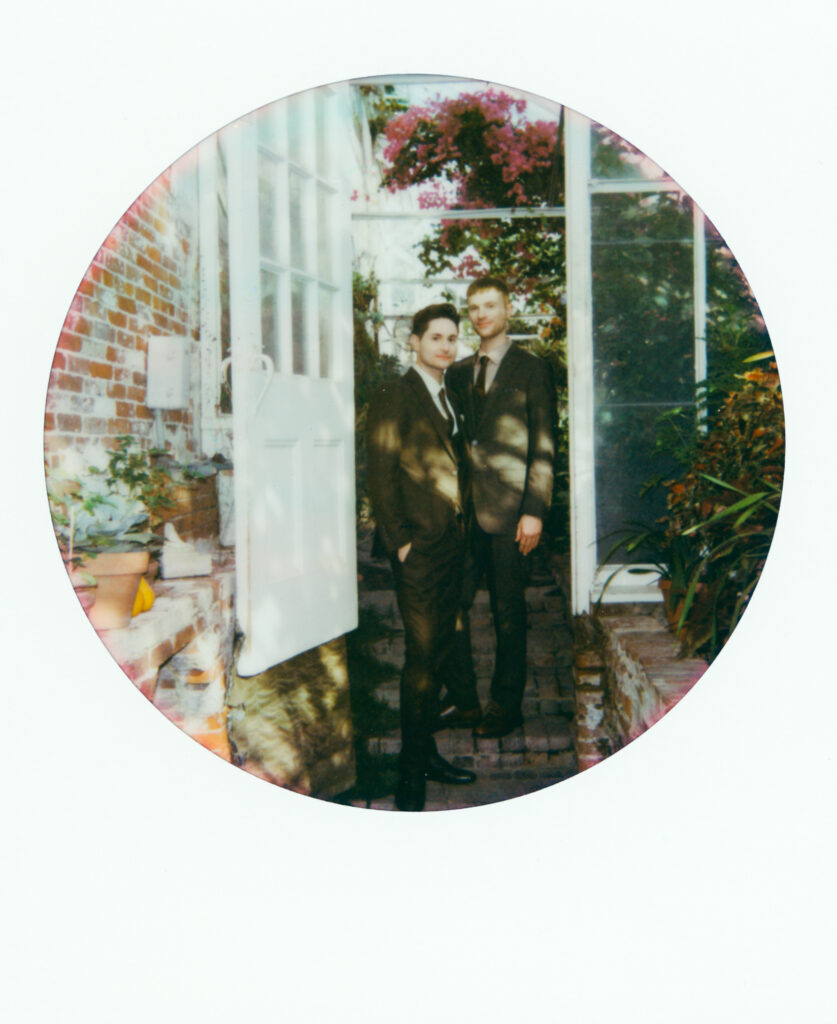 polaroid photo of queer couple posing in front of a green house