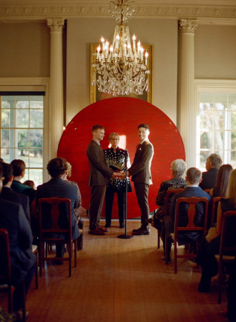 queer couple standing in front of a large red circular backdrop during wedding ceremony