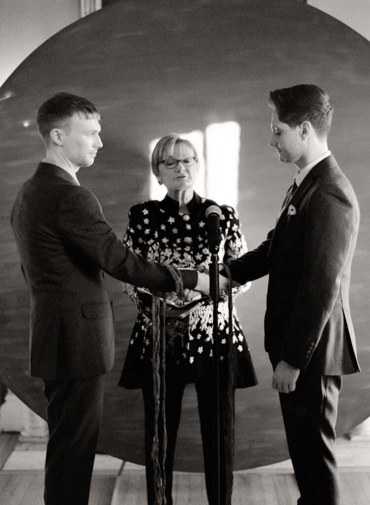 queer couple standing in front of a large red circular backdrop during wedding ceremony in black and white photo