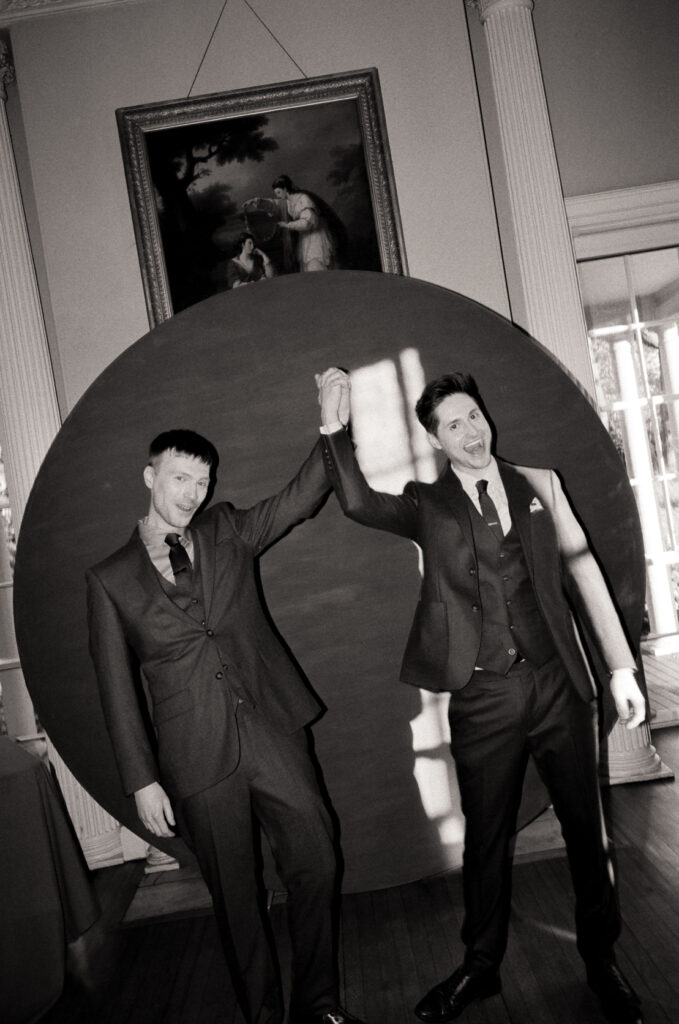 queer couple standing in front of a large red circular backdrop during wedding ceremony in black and white photo