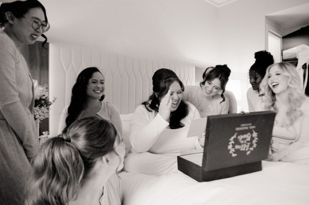 black and white film photograph of bride and bridesmaids sitting on bed reading letters from each other and laughing
