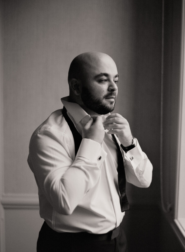 black and white film photo of groom buttoning his shirt before wedding