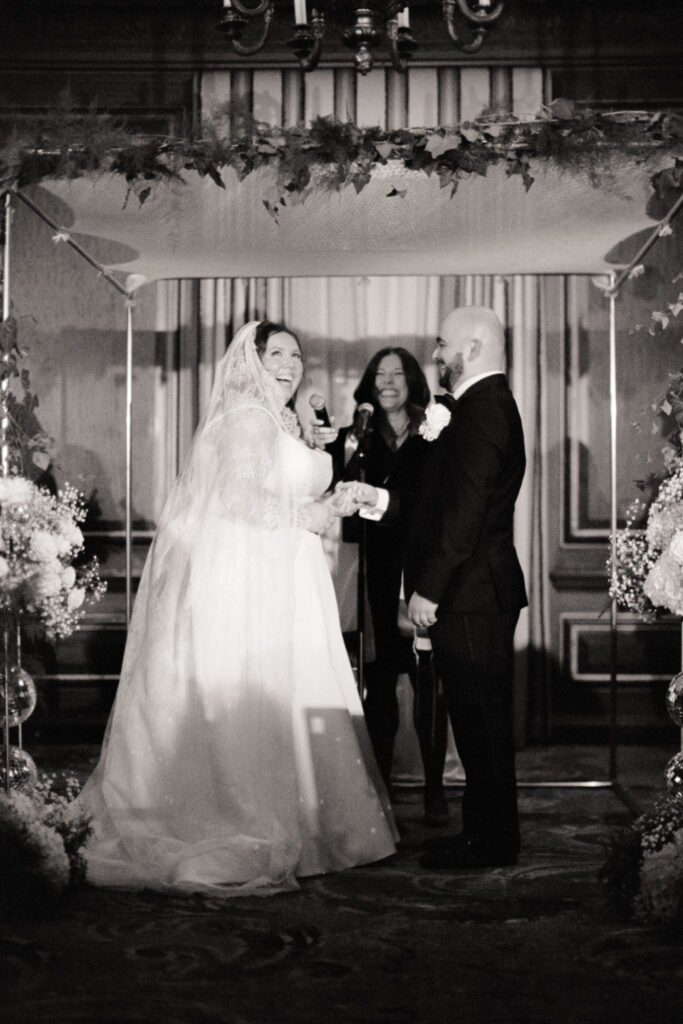 bride smiling and groom smiling at her during wedding ceremony under huppah