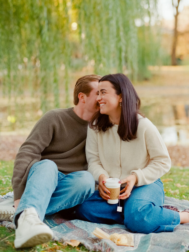 man whispering in women's ear who is holding coffee in a park 