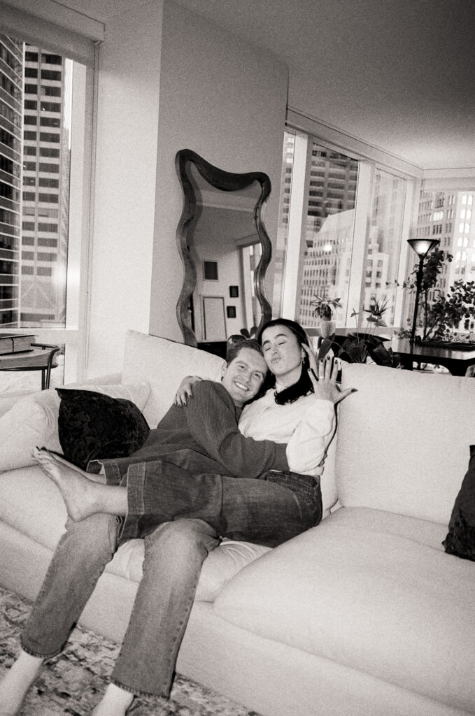 couple sitting on the couch and smiling holding up wedding ring  in black and white photo during film engagement photos 