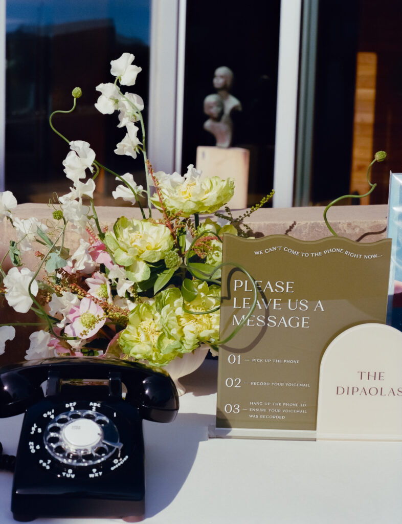 a dial phone with a sign that says leave us a message with floral arrangement behind it 