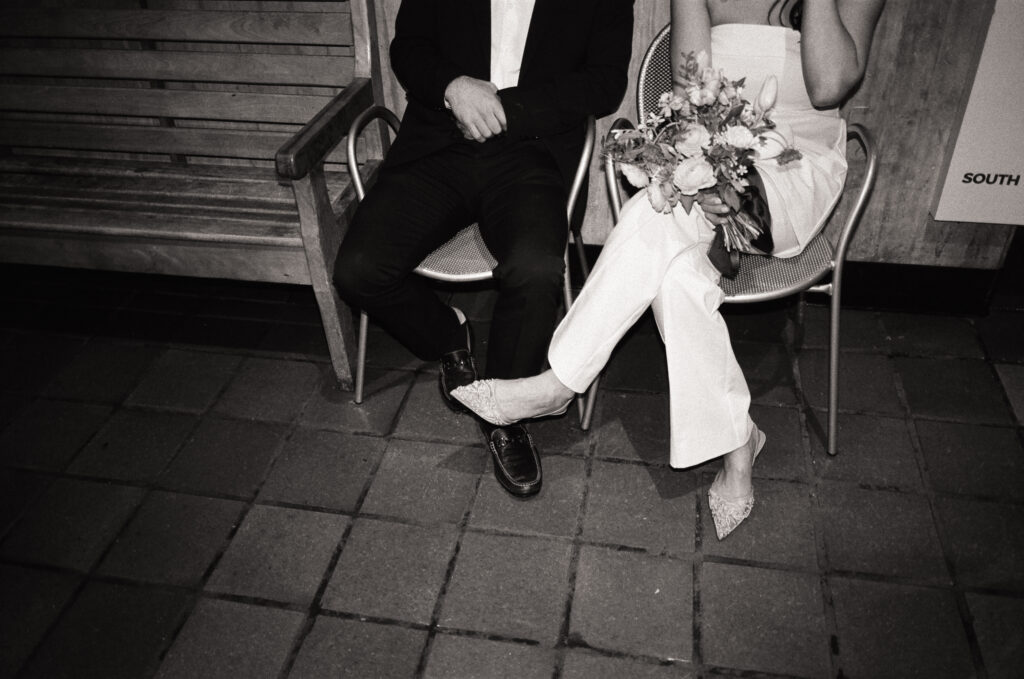 man in a suite sitting in a chair next to a woman in a white outfit holding a bouquet during a Boston City Hall elopement