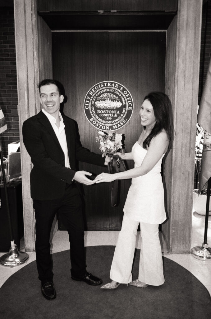 Man and woman standing in the Boston City Hall holding hands and smiling while saying their vows
