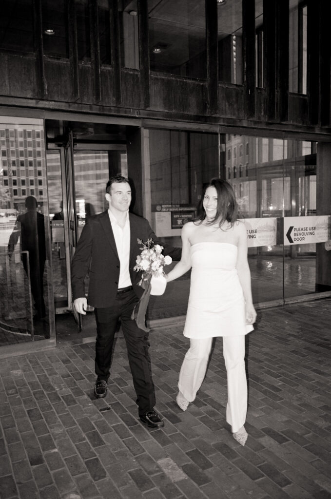 A Woman and a man walking out of city hall holding a wedding bouquet after eloping 