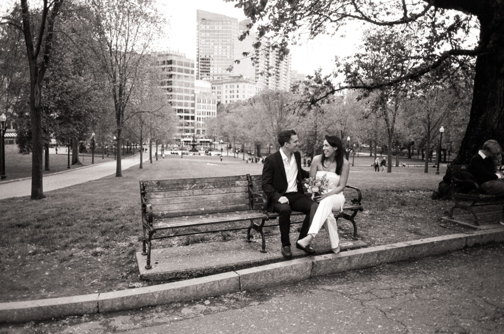 bride and groom sitting on a bench in a park smiling at one another in black and white photo 