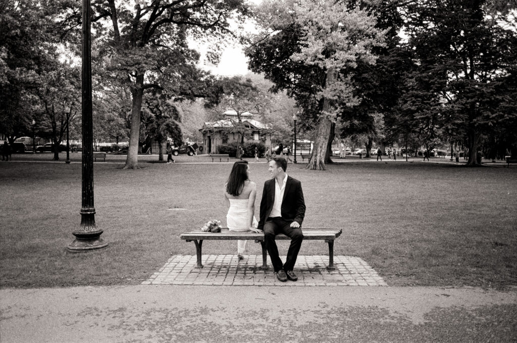 bride and groom sitting on a park bench looking at one another in black and white photo 