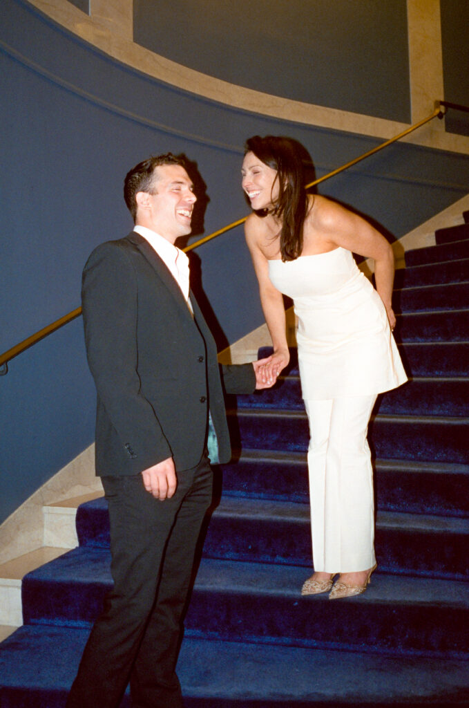 bride and groom standing on velvet blue stairs holding hands and laughing 