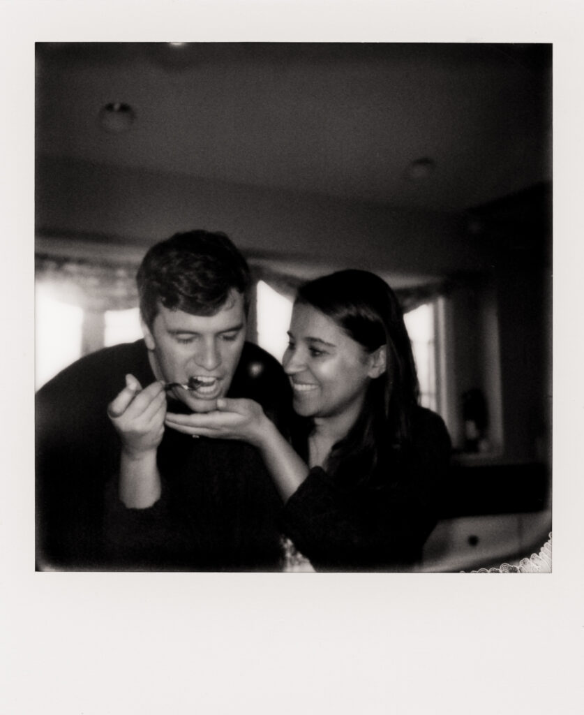 polaroid photo of woman holding a spoon up to a mans mouth, feeding him clams