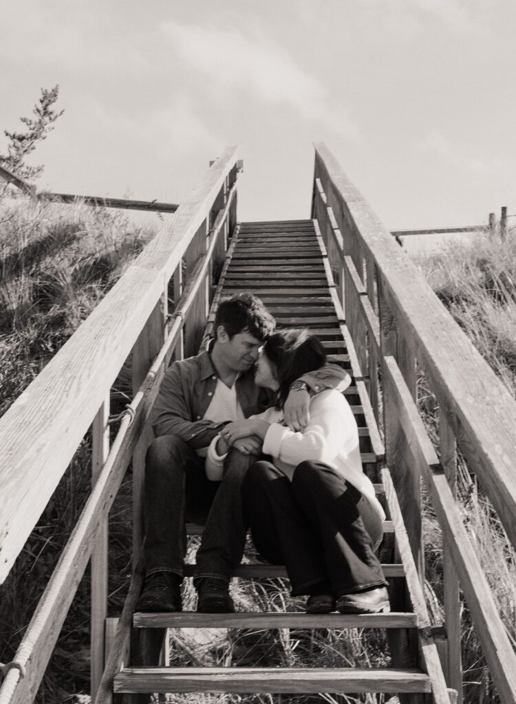 couple sitting on stairs that lead to the beach cuddles up and leaning towards one another